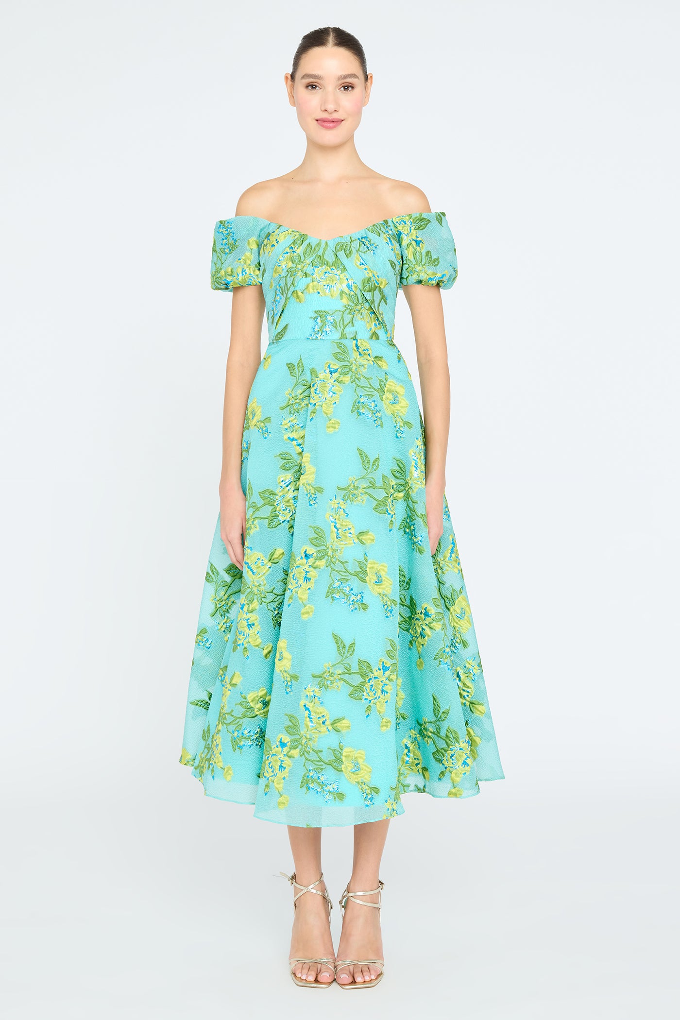 A woman wearing an off-shoulder mid-length dress with a floral pattern, in turquoise, green, and yellow colors.