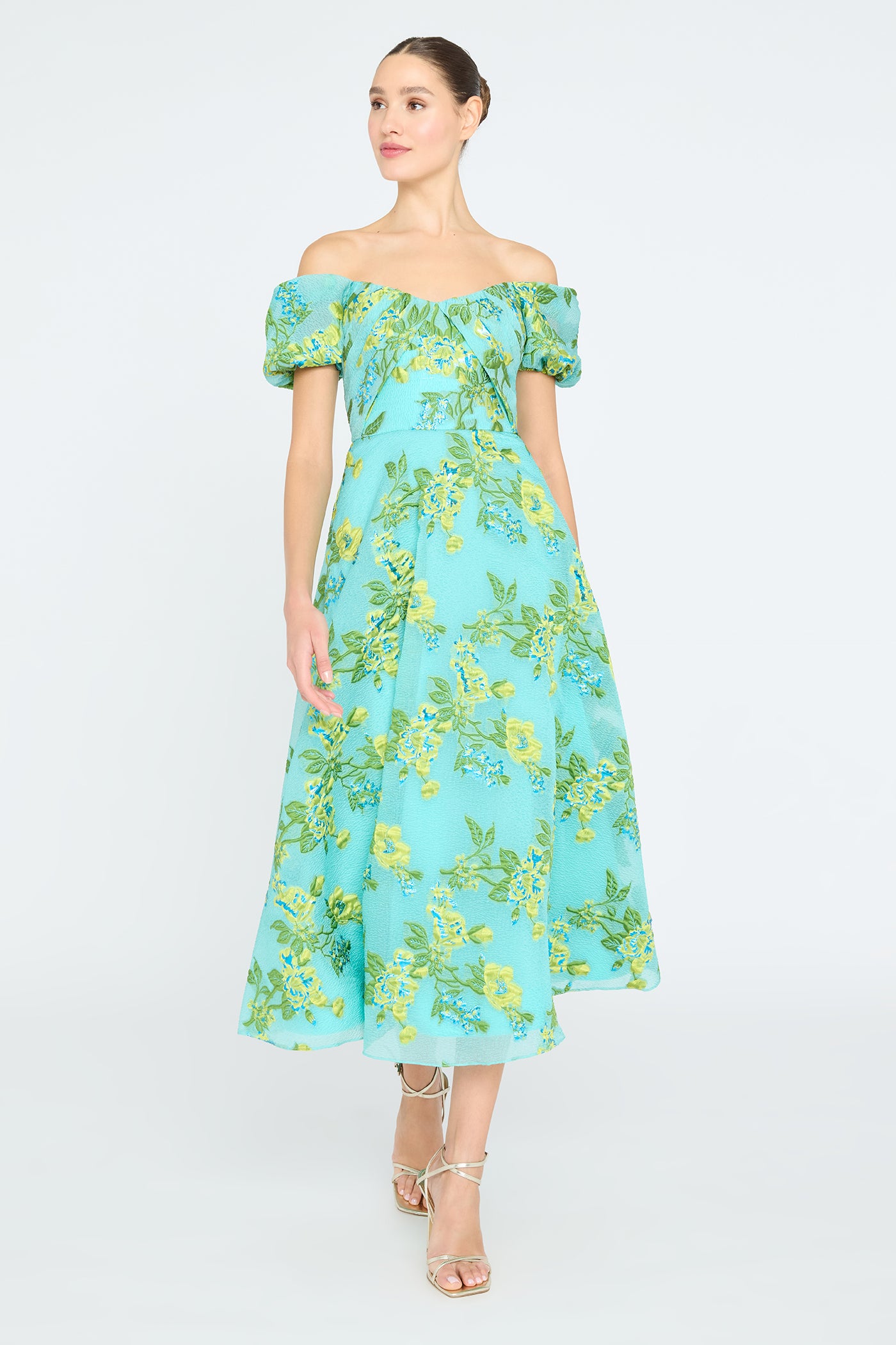 A woman wearing an off-shoulder mid-length dress with a floral pattern, in shades of green and blue, paired with beige accessories.